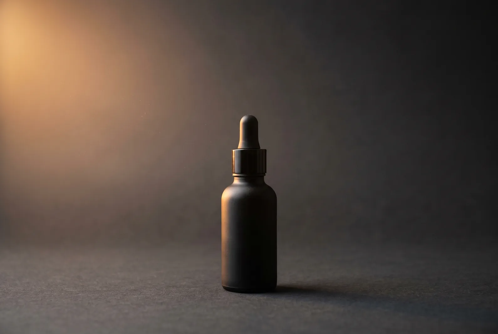 Hero packshot of a matte-black dropper bottle, warm amber rim light, near-black backdrop.